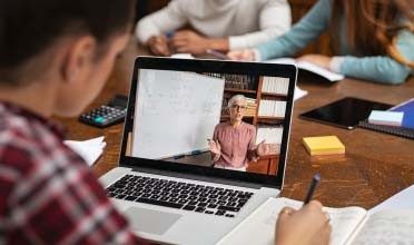 Image of a young student in a red and black plaid shirt looking at a laptop watching an online teacher give a class for Connections Academy.